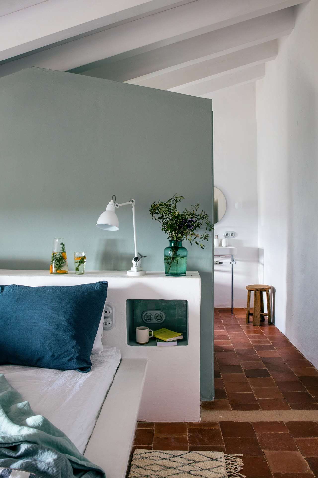 Es Bec d’Aguila — Es Bec d'Aguila, Balearic Islands, Spain — Minimalist bedroom with sage-green accent wall, white desk, brass desk lamp, terracotta tiles, exposed beam ceiling, and garden views through open passage.