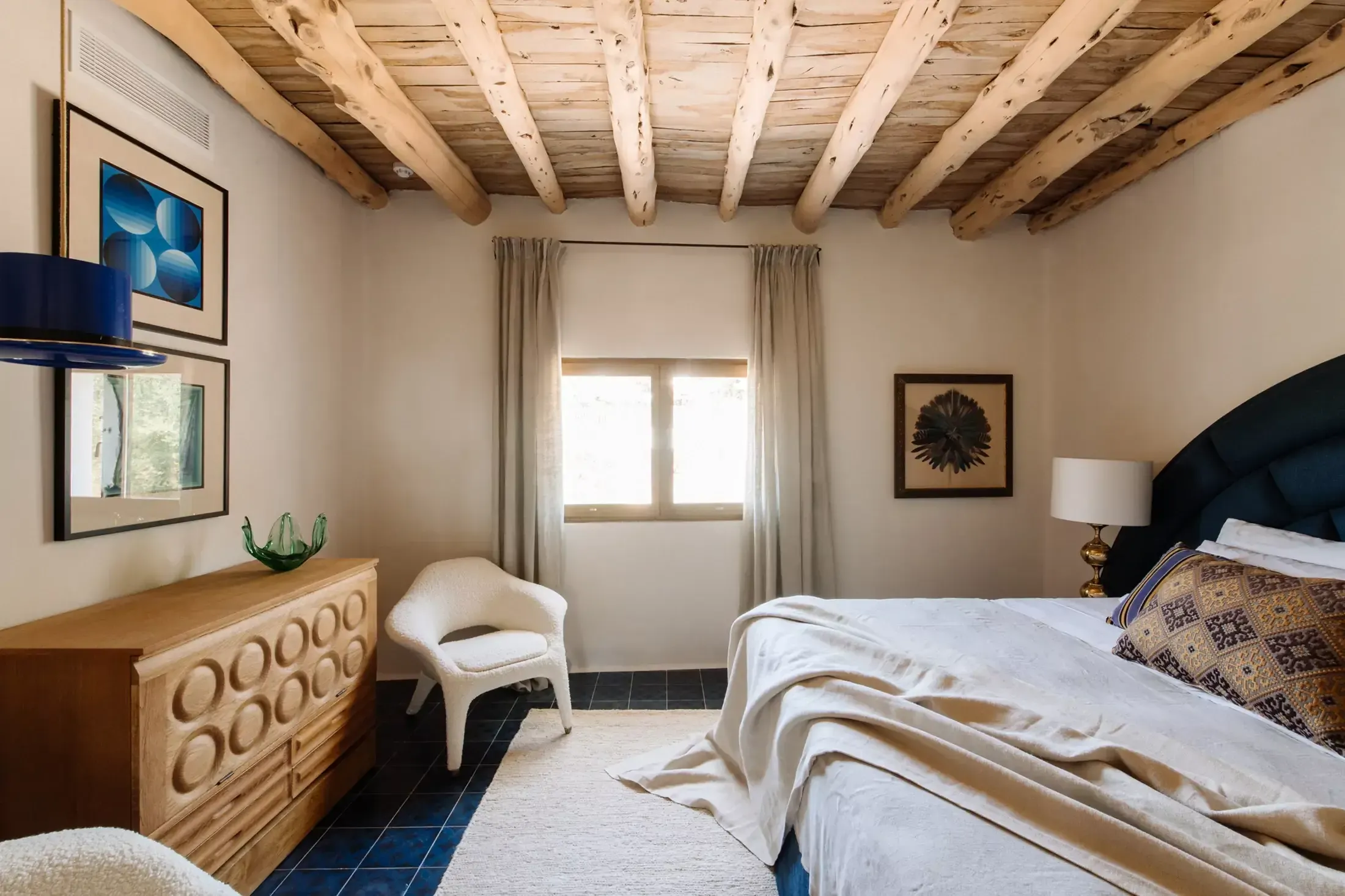 Finca La Inika, Balearic Islands, Spain — Master bedroom with exposed wooden ceiling beams, natural linen curtains, carved wood dresser, navy upholstered headboard, and slate flooring.