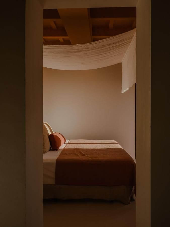 Amagatay, Balearic Islands — Minimalist bedroom with sheer fabric canopy, warm wood furnishings, and natural light through doorway.