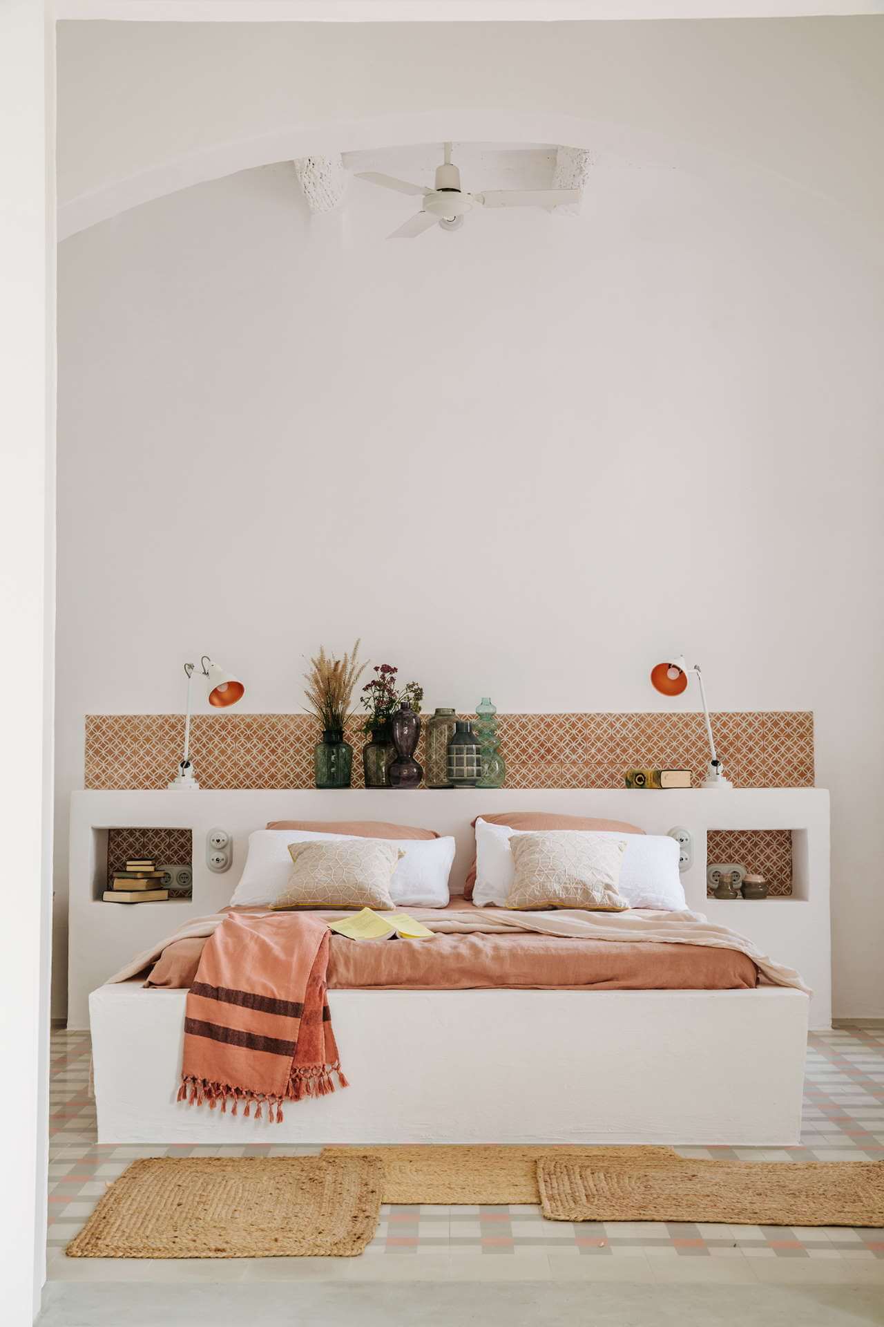 Es Bec d’Aguila — Es Bec d'Aguila, Balearic Islands, Spain — Minimalist bedroom with built-in white bed frame, geometric patterned headboard shelf, terracotta accents, natural fiber rugs, ceiling fan, and curated travel-worn decor.