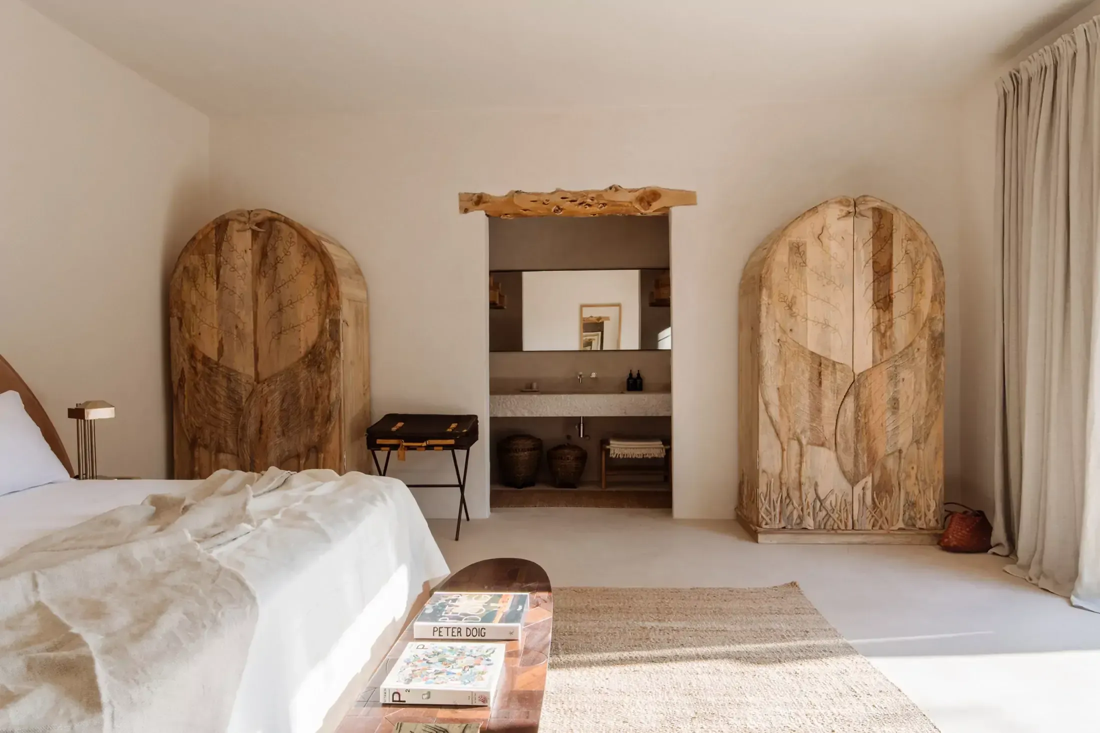 Finca La Inika, Balearic Islands, Spain — Minimalist bedroom with arched wooden doors, white linens, natural light, and carved wood wardrobe in neutral palette.