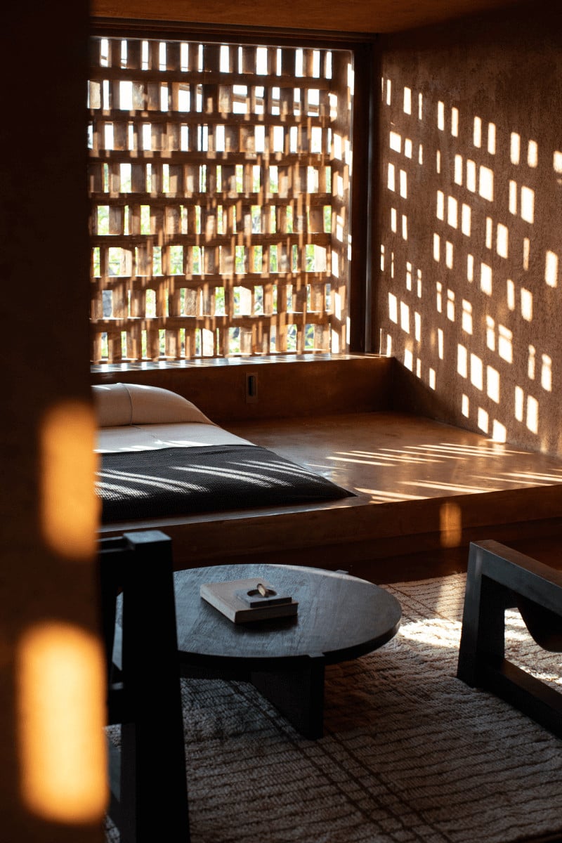 Further Hotel, Asia — Sunlit bedroom with latticed brick windows casting geometric shadows, minimalist wood bed frame, round stone table, warm ambient lighting.