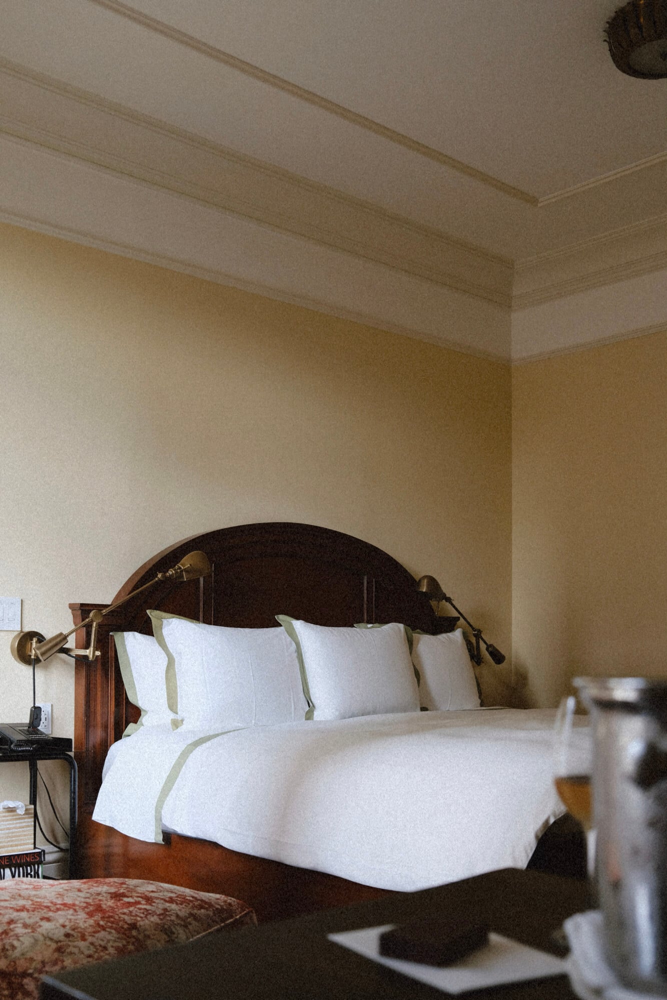 The Greenwich Hotel, North America — Elegantly appointed bedroom with dark wood bed frame, crisp white linens, curved headboard, brass sconces, cream walls, and coffered ceiling detail.
