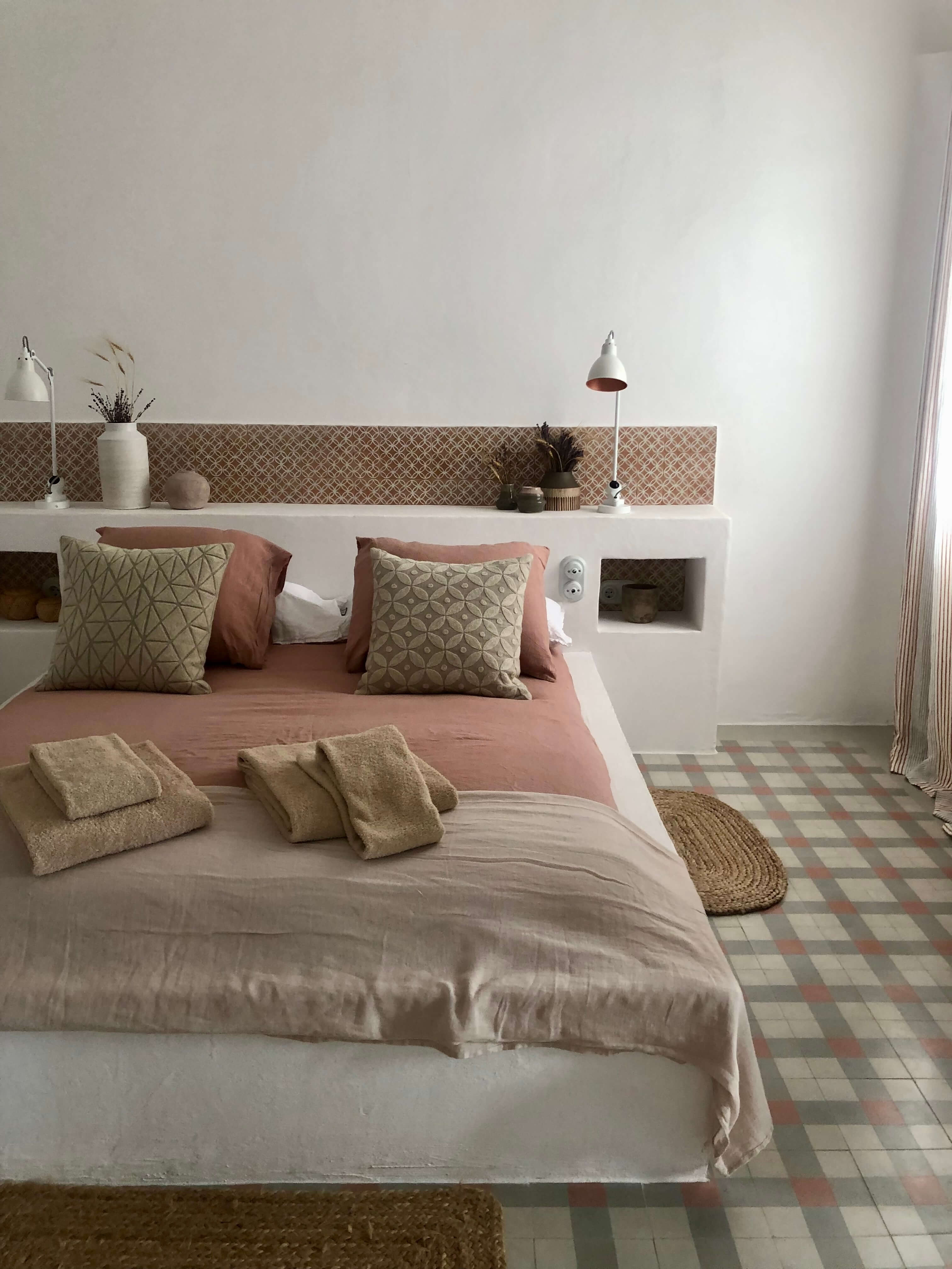 Es Bec d’Aguila — Es Bec d'Aguila, Balearic Islands, Spain — Minimalist bedroom with geometric patterned headboard, terracotta and neutral linens, pendant lamps, patterned tile flooring, and built-in shelving.