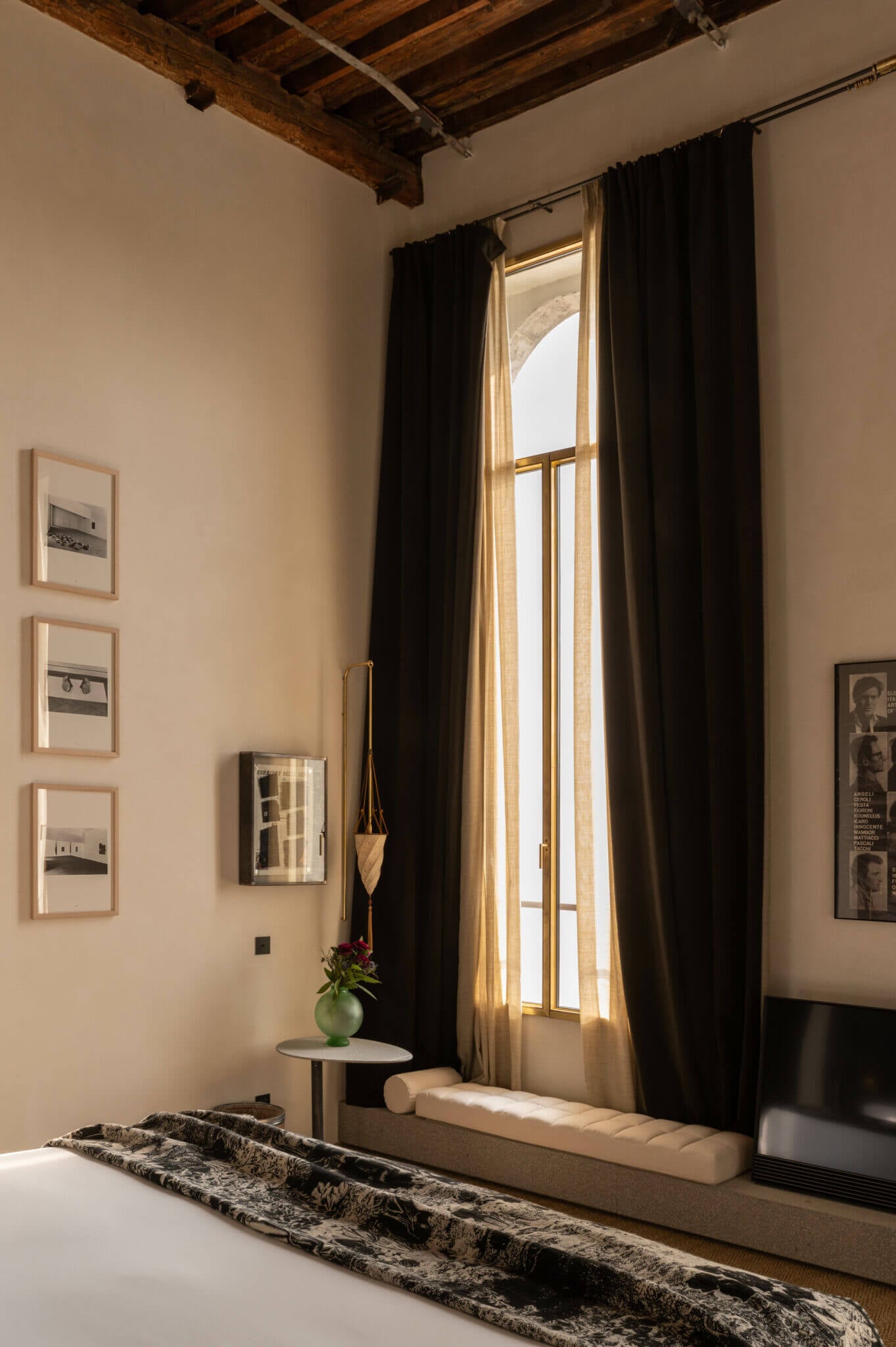The Venice Venice Hotel, Venice, Europe — Minimalist bedroom with exposed timber ceiling beams, tall shuttered windows, black curtains, gallery wall, and natural light streaming across neutral stone flooring.