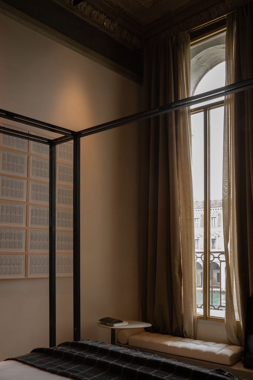 The Venice Venice Hotel, Venice, Italy — Minimalist bedroom with black metal canopy bed, beige linen, arched window overlooking canal, ornate ceiling detail, warm natural light.