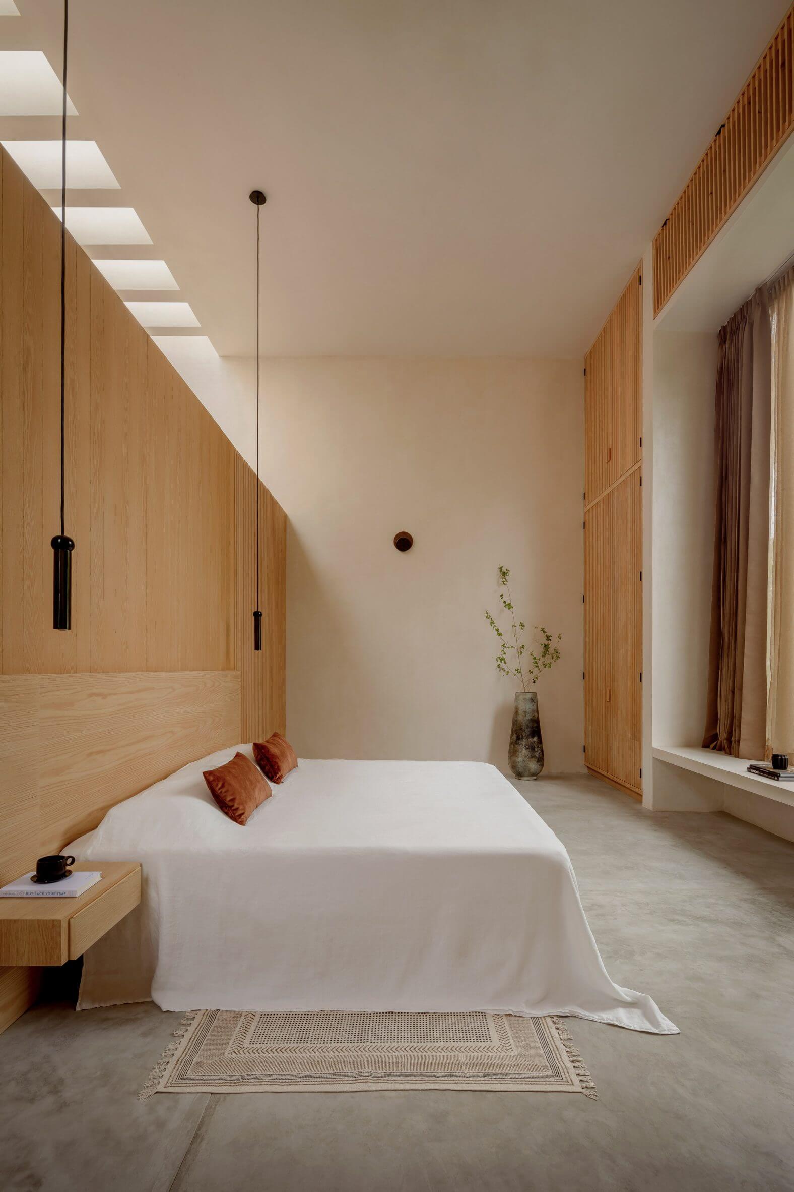 Villa Cava, Mexico — Minimalist bedroom with natural wood wall paneling, skylights, white bedding, pendant lights, and concrete flooring with geometric rug.