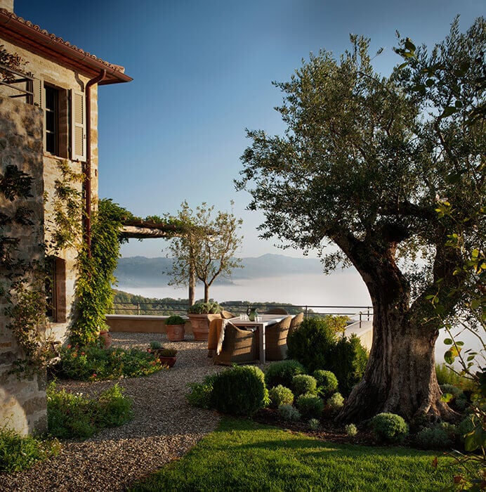 Castello di Reschio, Italy — Stone villa terrace with olive tree, wicker seating, and misty lake view under clear sky.