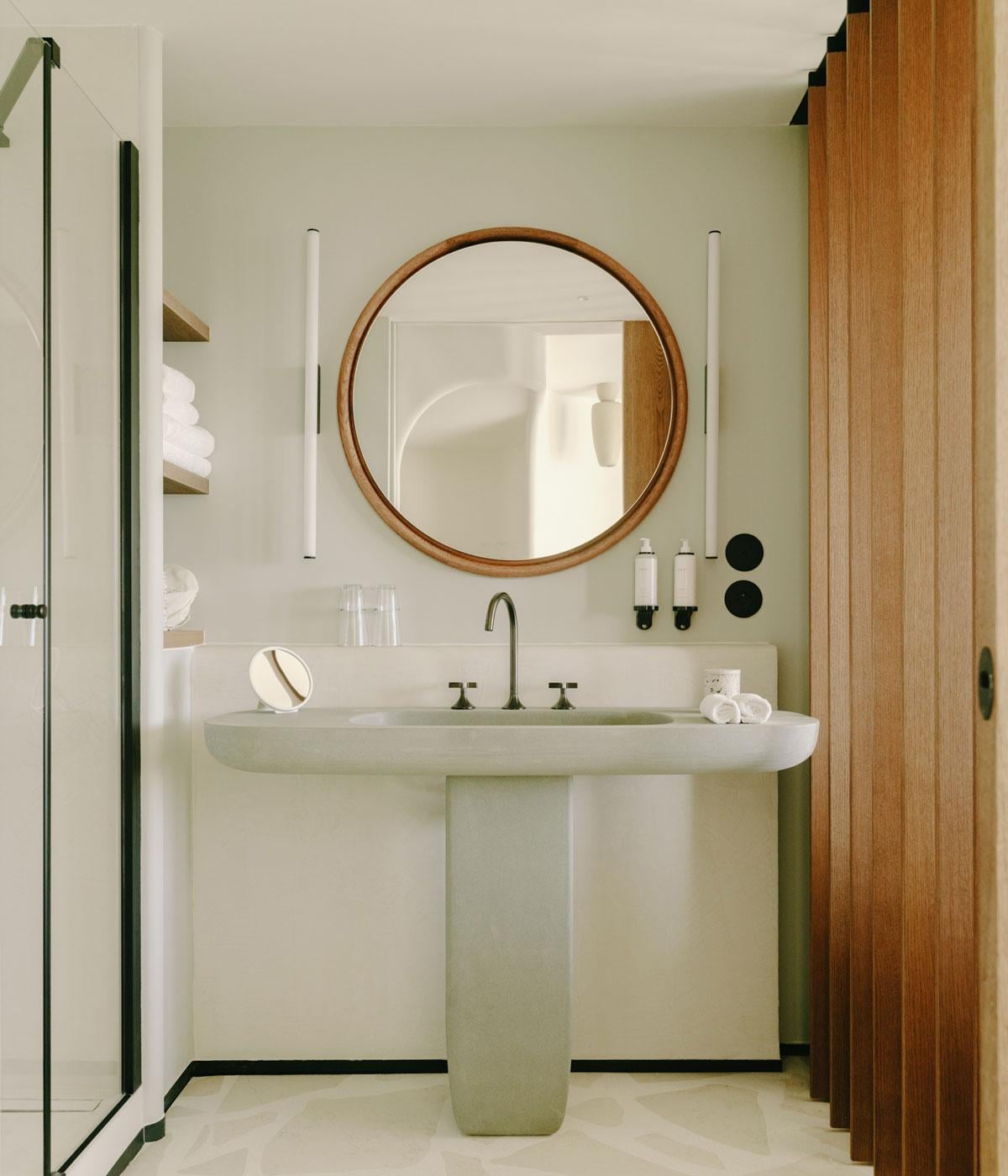 Belle Plage, Europe — Minimalist bathroom with pale green pedestal sink, round wooden mirror, black-framed shower enclosure, and warm wood accents.