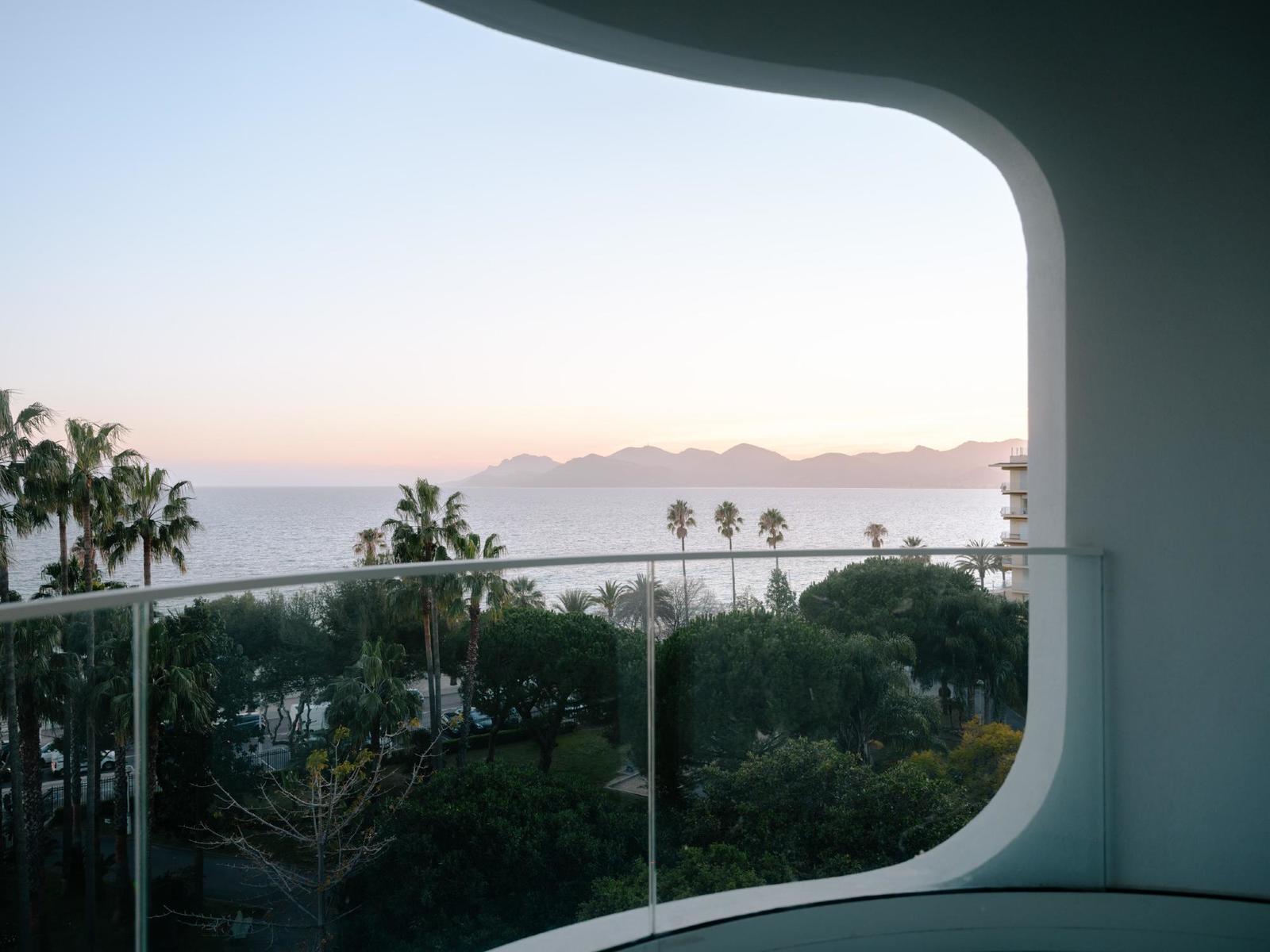 Belle Plage, Europe — Curved glass balcony framing Mediterranean seascape at sunset with palm trees and distant mountains.