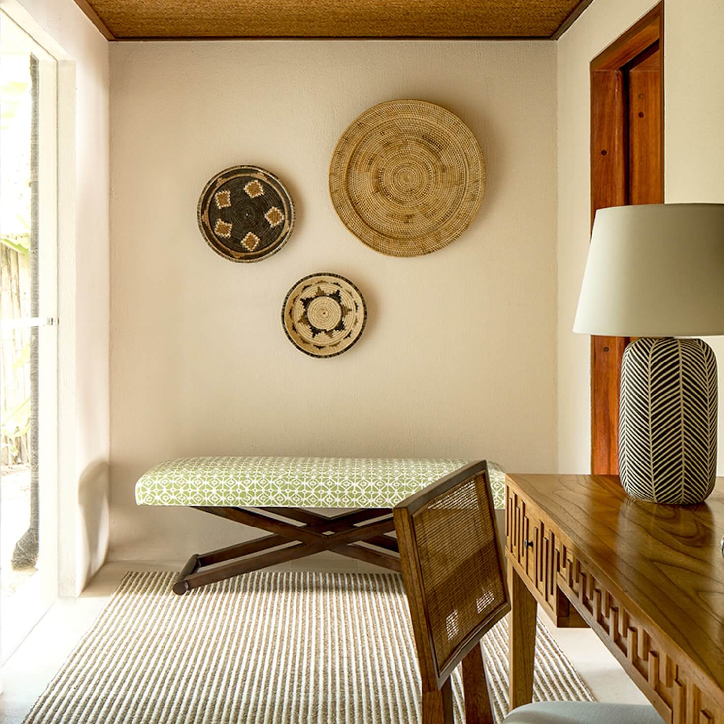 NIHI Sumba, Asia — Coastal retreat bedroom with woven basket wall art, wooden X-frame bench, striped linen rug, natural light, and minimalist organic décor.