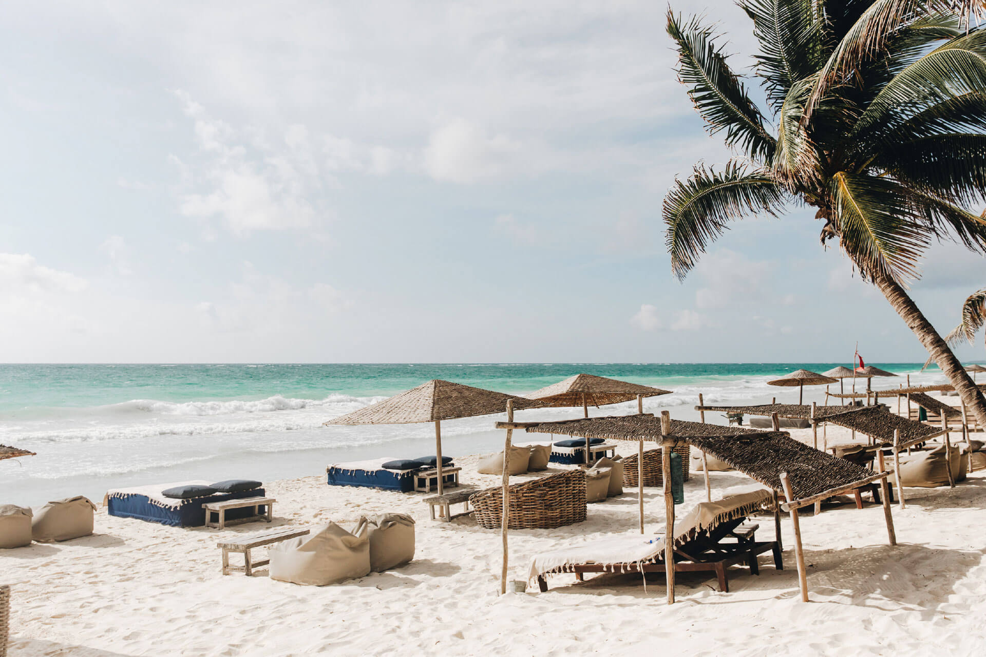 Be Tulum, Mexico — Beach club with woven umbrellas, natural wood loungers, wicker cabanas, and palm fronds overlooking turquoise Caribbean waters.