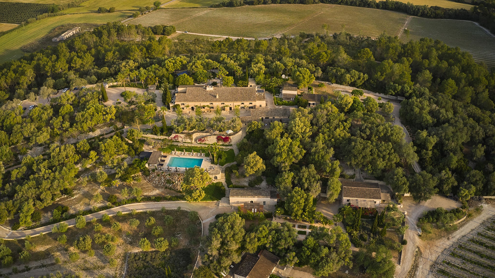 Gardens & Orchards, Balearic Islands, Spain — Stone manor nestled in cypress woodland with terraced gardens, swimming pool, and cultivated fields beyond, golden hour light.