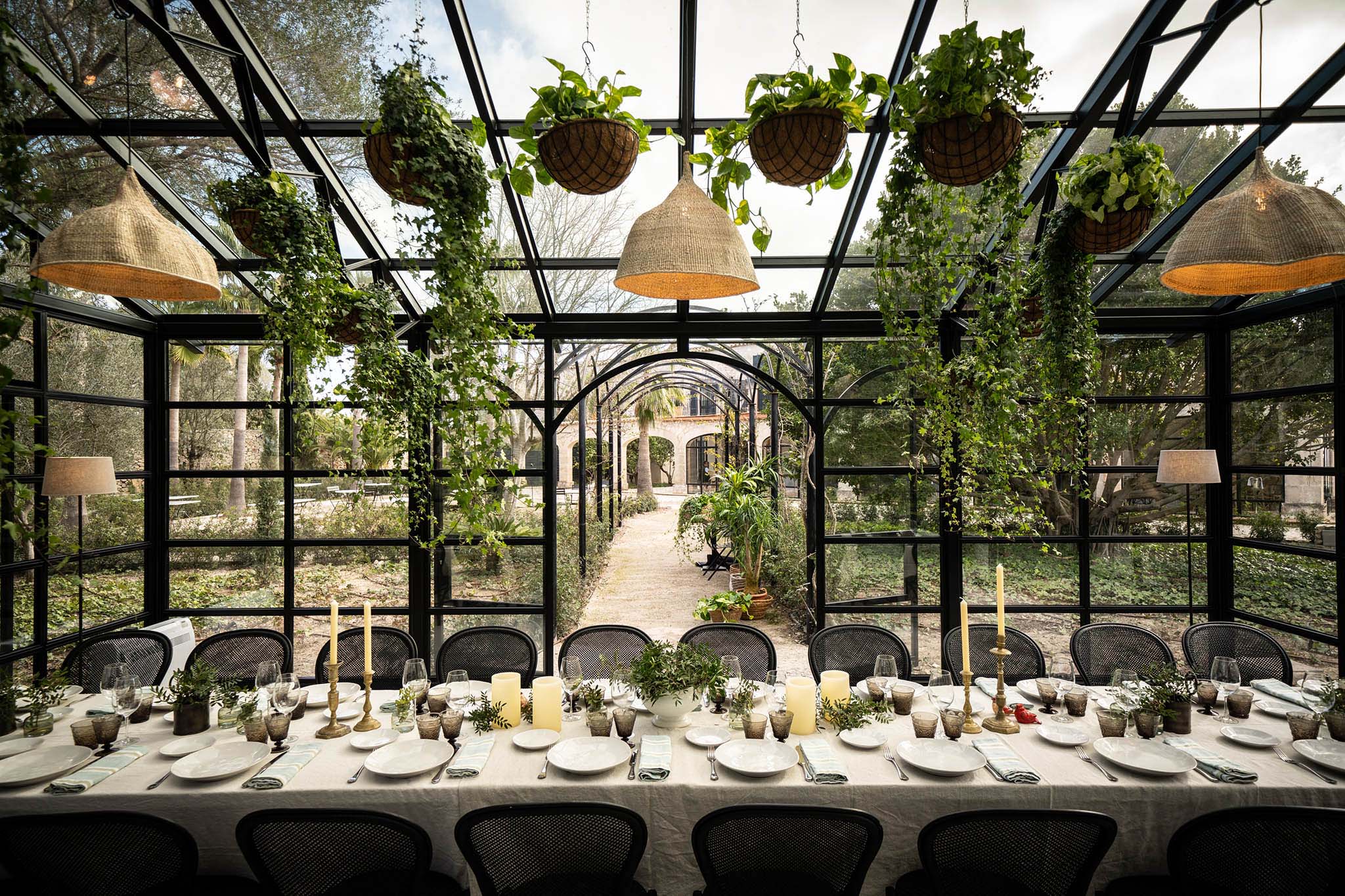 Finca Serena, Balearic Islands, Spain — Greenhouse dining room with black steel framework, hanging plants, woven pendant lights, and garden views through expansive glazing.