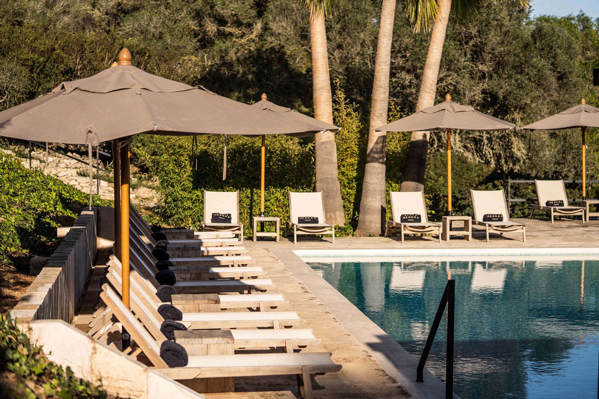 Finca Serena, Balearic Islands, Spain — Poolside lounge with taupe umbrellas, teak chairs, palm trees, and natural stone terracing in Mediterranean luxury setting.