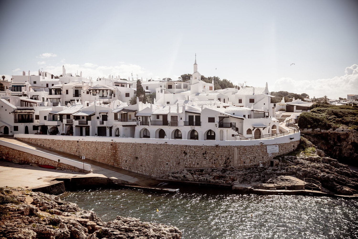 Binibeca, Balearic Islands, Spain — Whitewashed Mediterranean village with arched waterfront buildings, stone walls, and turquoise cove, afternoon light.