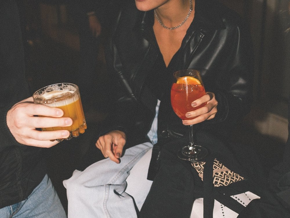 Birdman, Europe — Two people holding cocktails at an evening gathering, wearing black leather jacket and light blue denim, ambient indoor lighting.