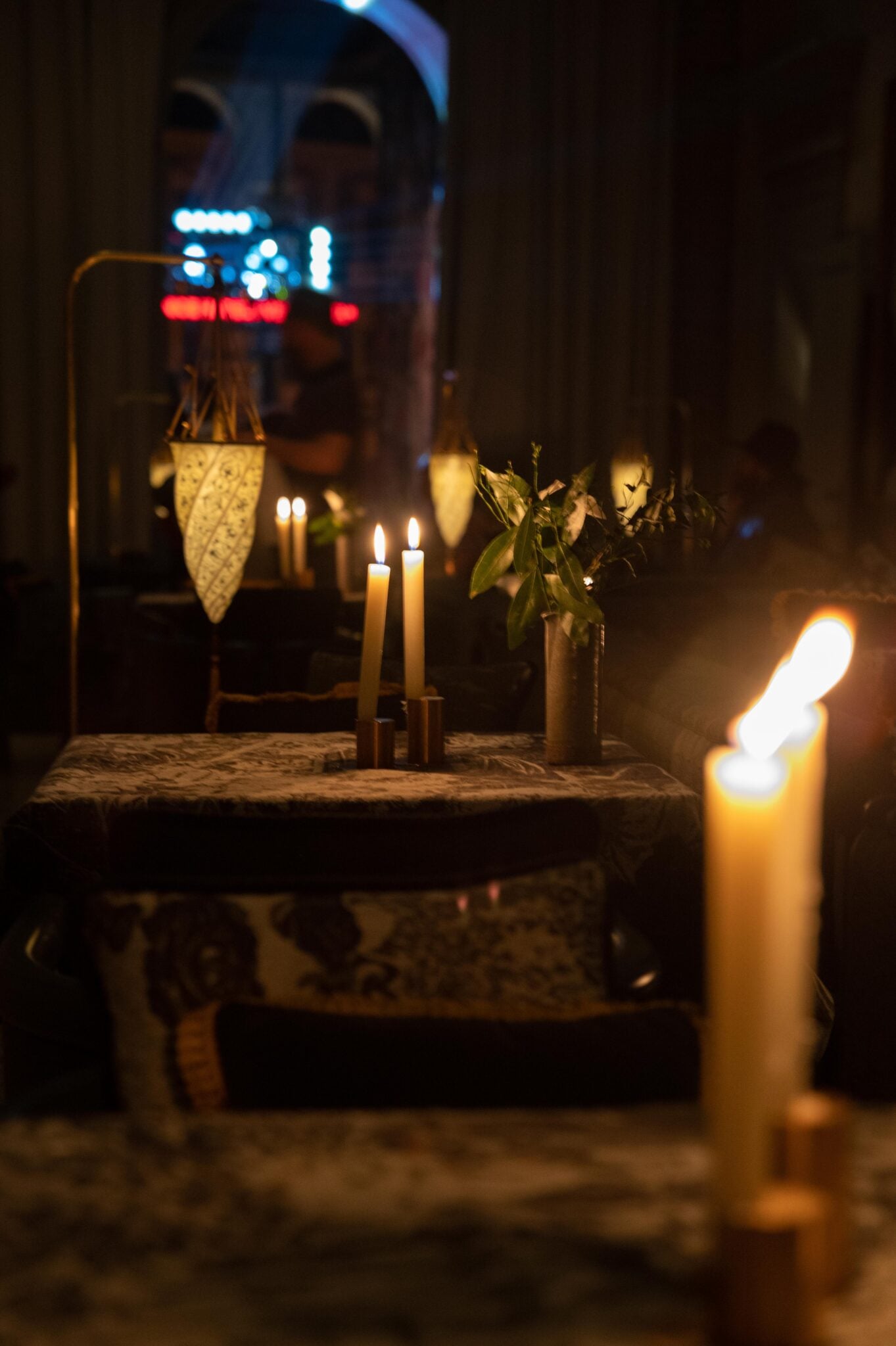 The Venice Venice Hotel, Europe — Candlelit table with fresh flowers and brass candelabra, neon-lit storefront visible through window at night.