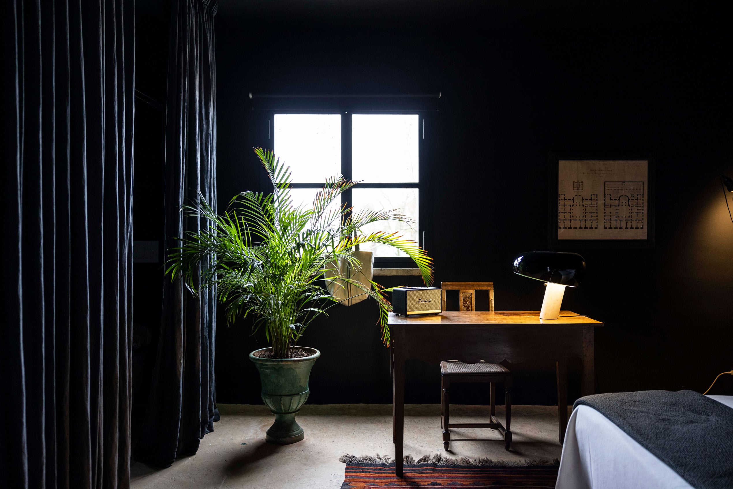 Palazzo Daniele, Europe — Dark bedroom with tall windows, potted palm, wooden desk, architectural drawing, and dramatic curtains creating sophisticated minimalist ambiance.