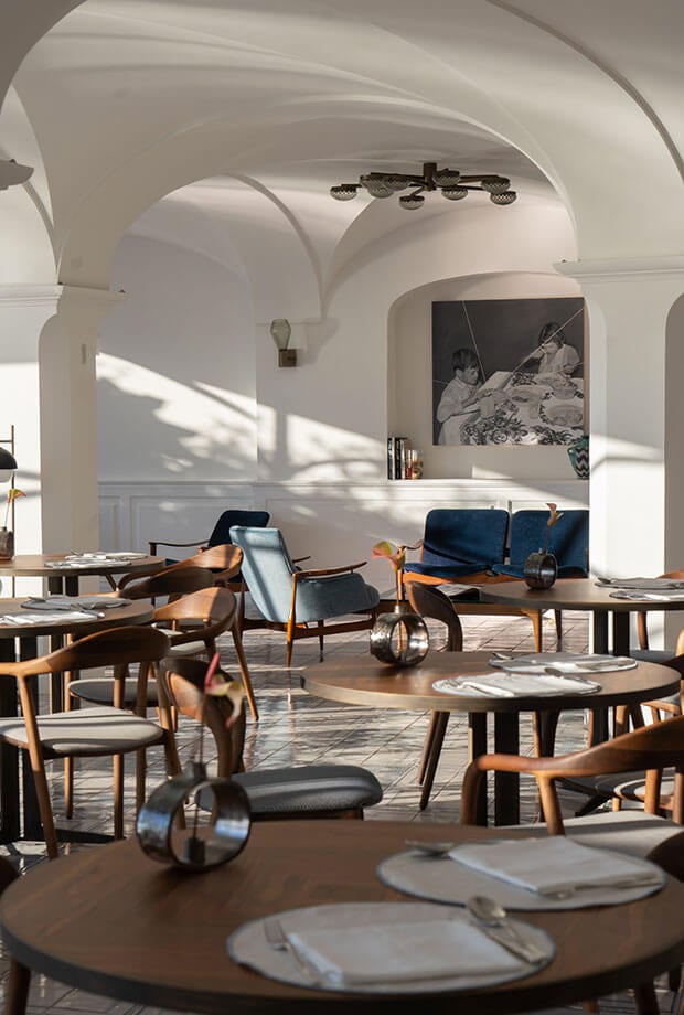 Alici, Europe — Arched white interiors with natural light, mid-century wooden furniture, navy upholstery, and round tables set for dining in minimalist luxury hospitality space.