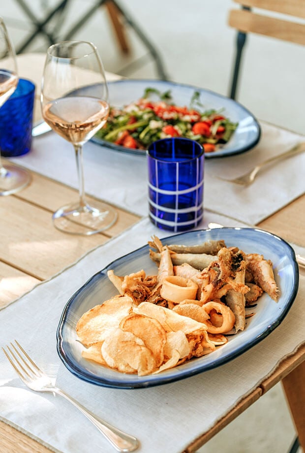 Beach Restaurant & Bar, Europe — Elegant seafood platter with crispy calamari and fish chips, rosé wine, and fresh vegetable salad on blue-rimmed ceramic plates, natural light dining setup.