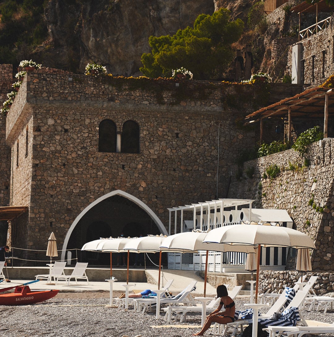 Borgo Santandrea, Europe — Beachfront stone architecture with arched entrance, white loungers and canvas canopies, Mediterranean coastal setting.