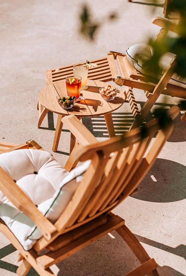 Borgo Santandrea, Europe — Wooden deck chairs with white cushions beside round side table holding cocktail and olives, dappled sunlight casting striped shadows on terracotta floor.