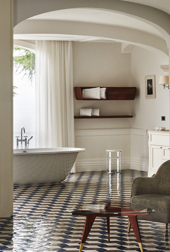 Borgo Santandrea, Europe — Spa bathroom with curved archway, freestanding tub, geometric floor tiles, dark wood shelving, cream palette, and natural light from arched window.
