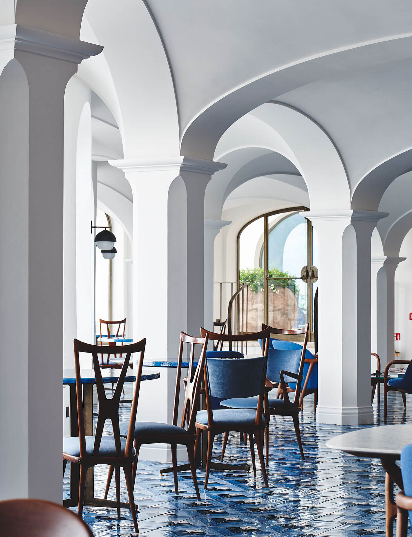 La Libreria Gourmet Restaurant, Europe — Mediterranean colonnade with arched ceilings, blue tile flooring, mid-century wooden chairs, and sea-view terrace beyond glass doors.