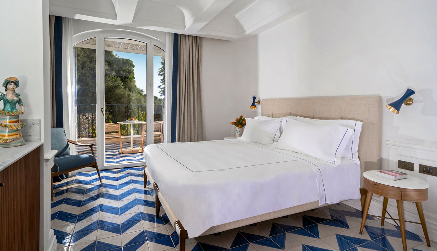 Borgo Santandrea, Europe — Bright bedroom with beige upholstered bed, geometric blue-striped flooring, arched glass doors opening to terrace with garden views, mid-century modern furnishings.