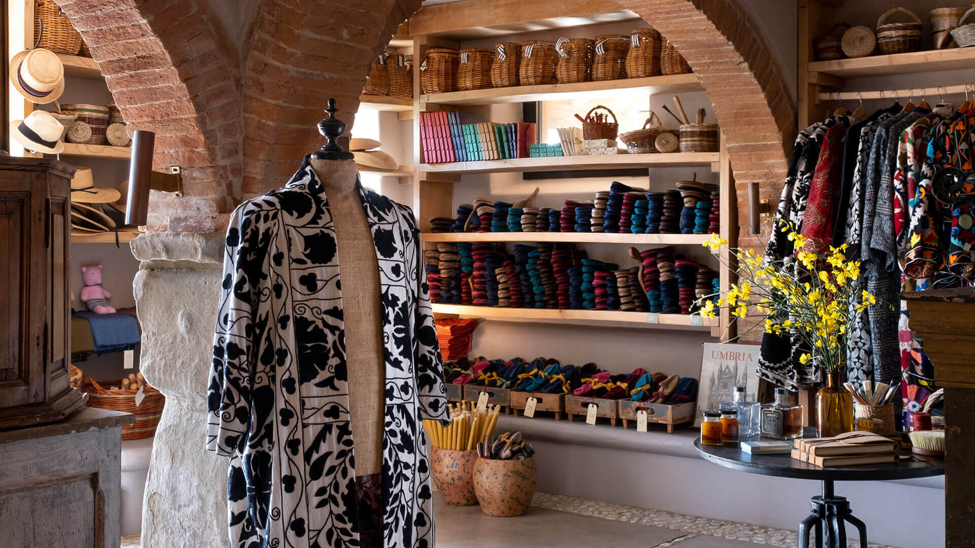 Bottega di Reschio, Umbria, Italy — Artisan textile boutique with brick archways, curated fabric displays, patterned scarves on dress form, and natural light illuminating shelved materials.