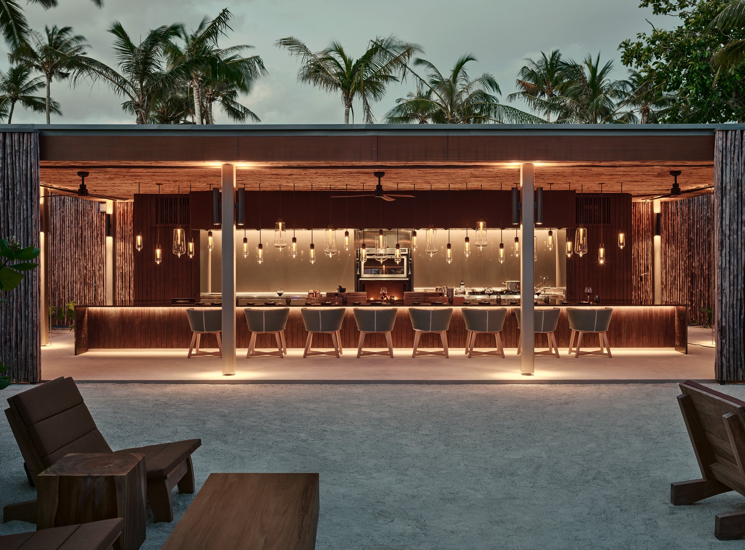 Brasa, Asia — Illuminated timber bar pavilion with pendant lighting, open-air seating, and palm-lined tropical setting at dusk.