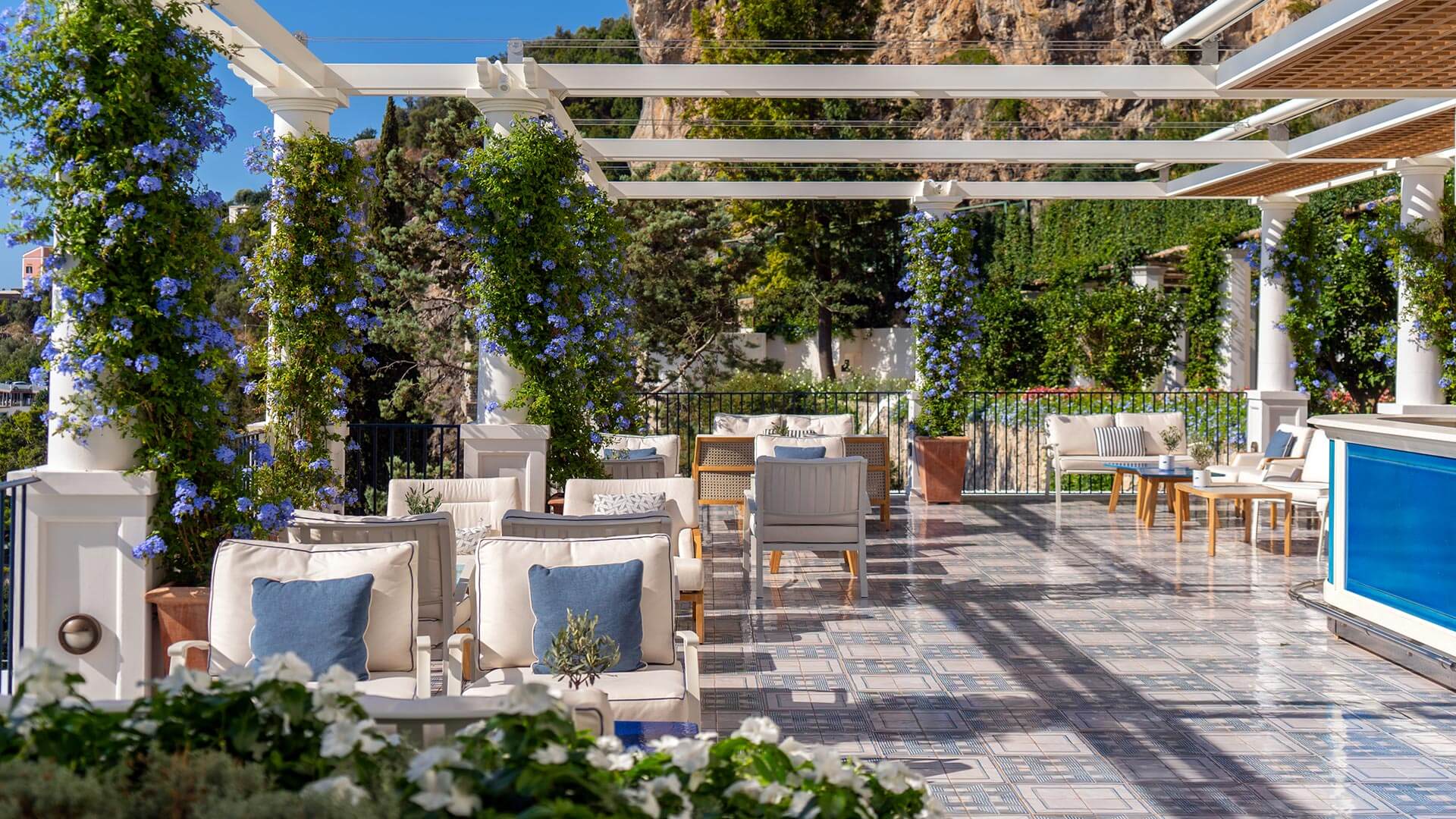 La Terrazza Bar, Europe — Pergola-shaded terrace with blue plumbago vines, cream lounge seating, stone paving, and cliff backdrop.