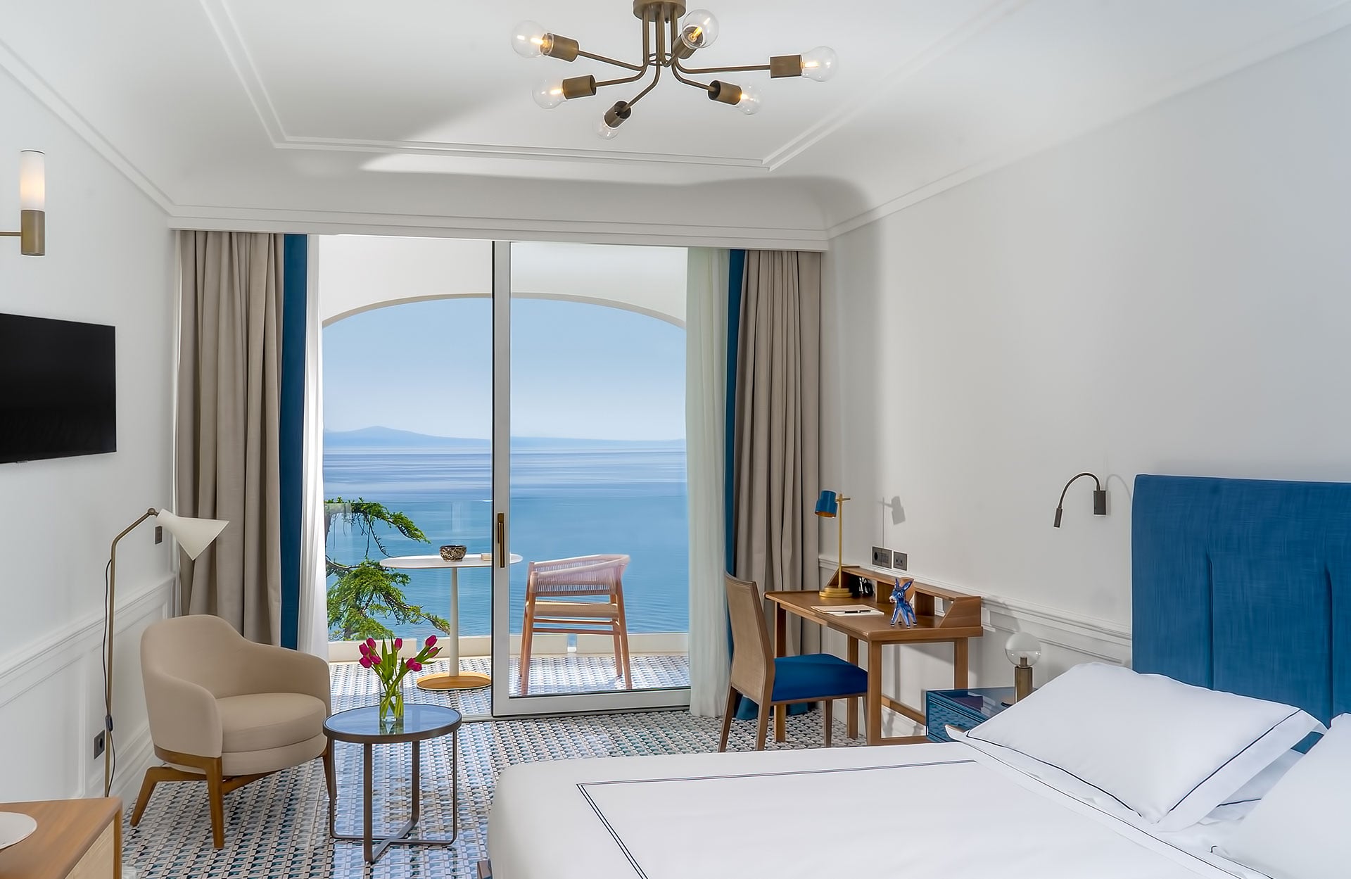 Borgo Santandrea, Europe — Luxury beachfront suite with arched glass doors, sea views, blue velvet headboard, mid-century furniture, and minimalist coastal design.