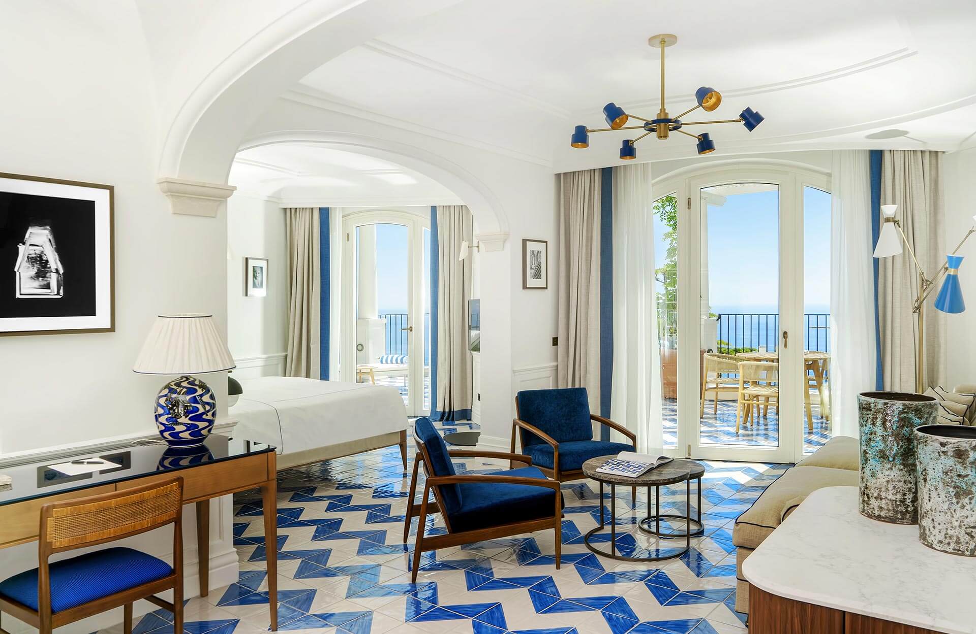 Borgo Santandrea, Europe — Coastal suite with arched ceilings, blue velvet chairs, geometric floor tiles, and sea views through French doors opening to balcony.
