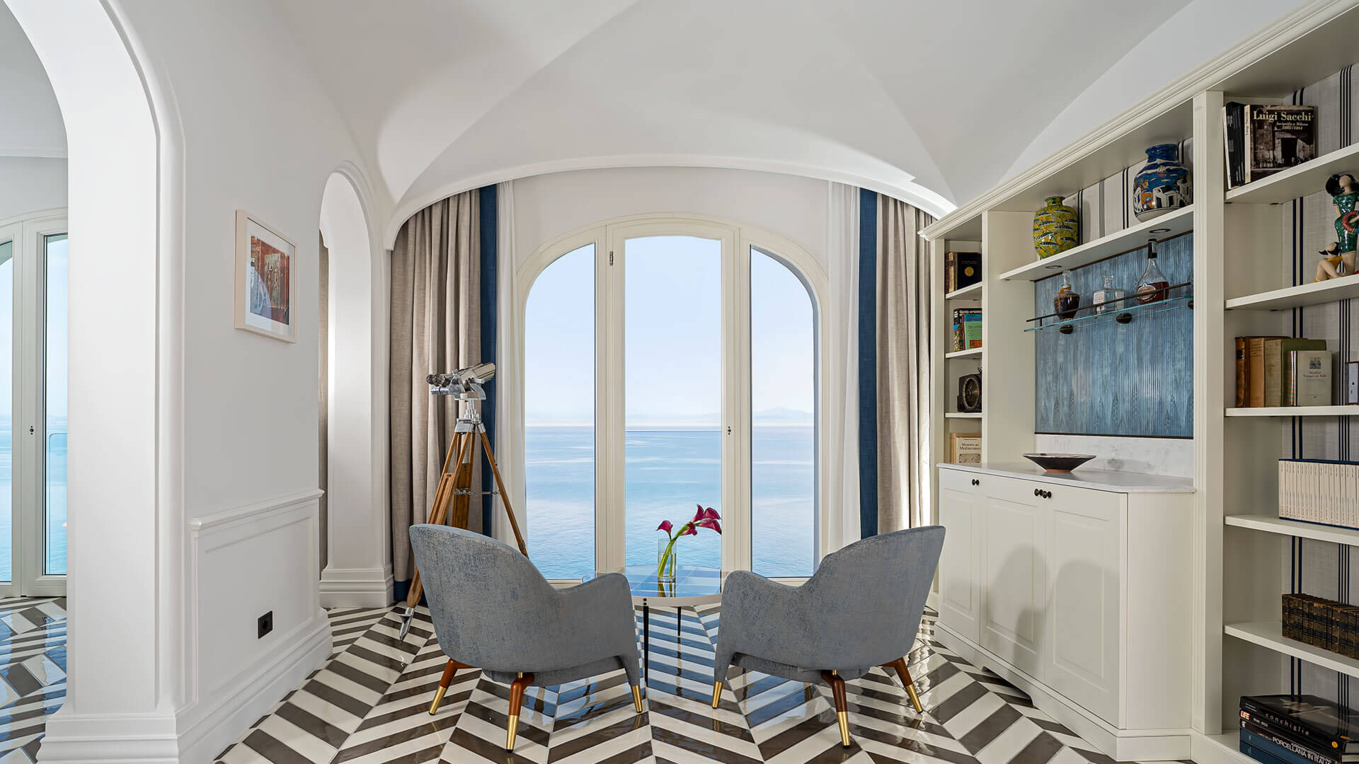 Borgo Santandrea, Europe — Coastal suite with arched barrel ceiling, curved French doors opening to sea view, striped marble floor, built-in shelving, and modern furnishings.