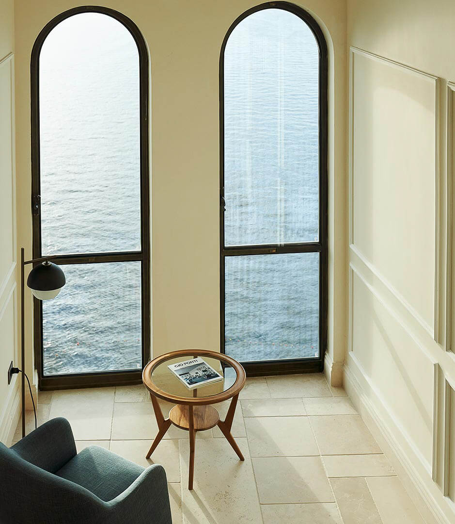 Borgo Santandrea, Europe — Arched black-frame doors open to sea views from minimalist room with wooden side table and upholstered chair.