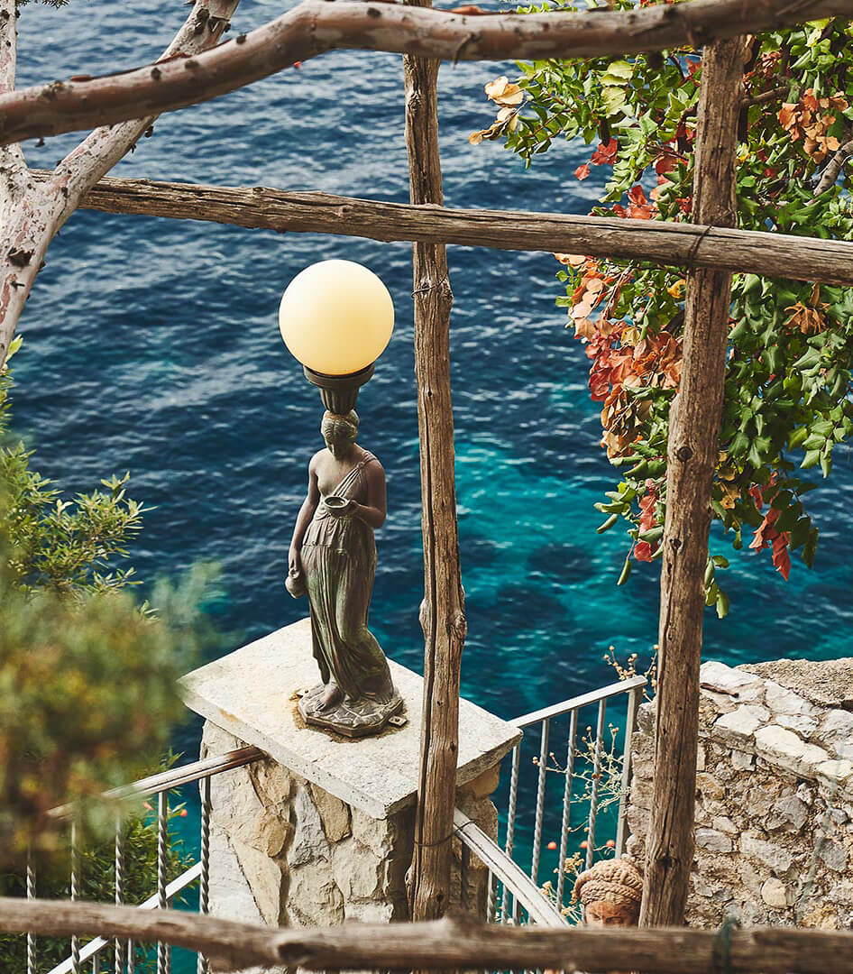 Borgo Santandrea, Europe — Bronze female statue with glowing orb beneath wooden pergola, overlooking turquoise Mediterranean sea with ivy-draped stone terrace.