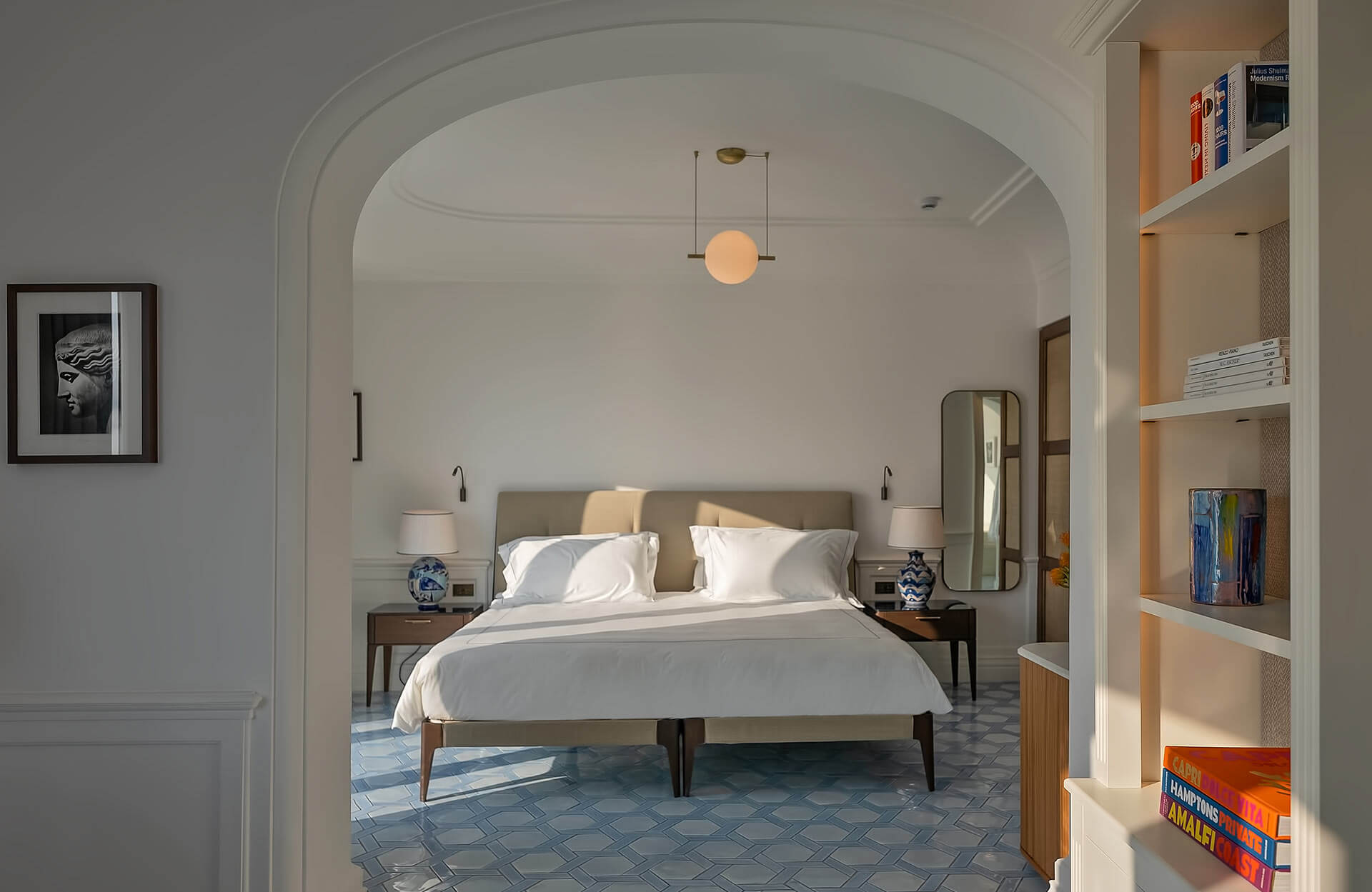 Borgo Santandrea, Europe — Modern bedroom with arched doorway, neutral palette, natural light, blue geometric floor tiles, mid-century furnishings, floating pendant light, built-in shelving.
