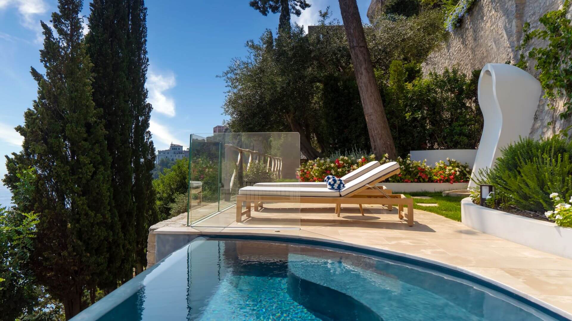 Borgo Santandrea, Europe — Infinity pool with travertine deck, cypress trees, and limestone terrace overlooking Mediterranean coast with modernist loungers and sculptural white furniture.