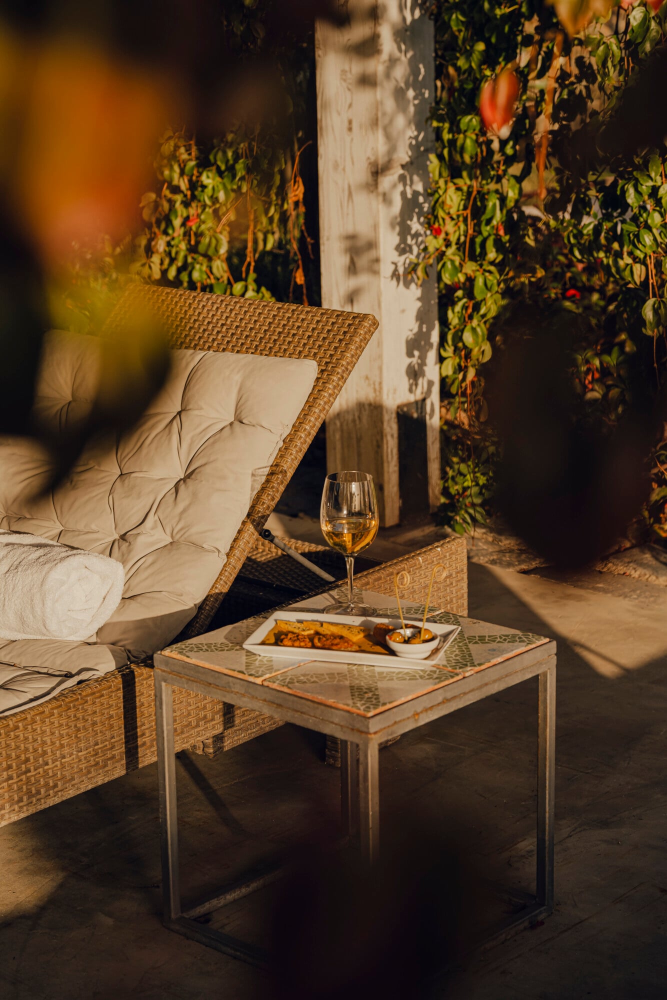 Susafa, Italy — Woven lounge chair with cream cushions beside side table holding wine glass and plate, ivy-covered whitewashed wall and golden afternoon light.