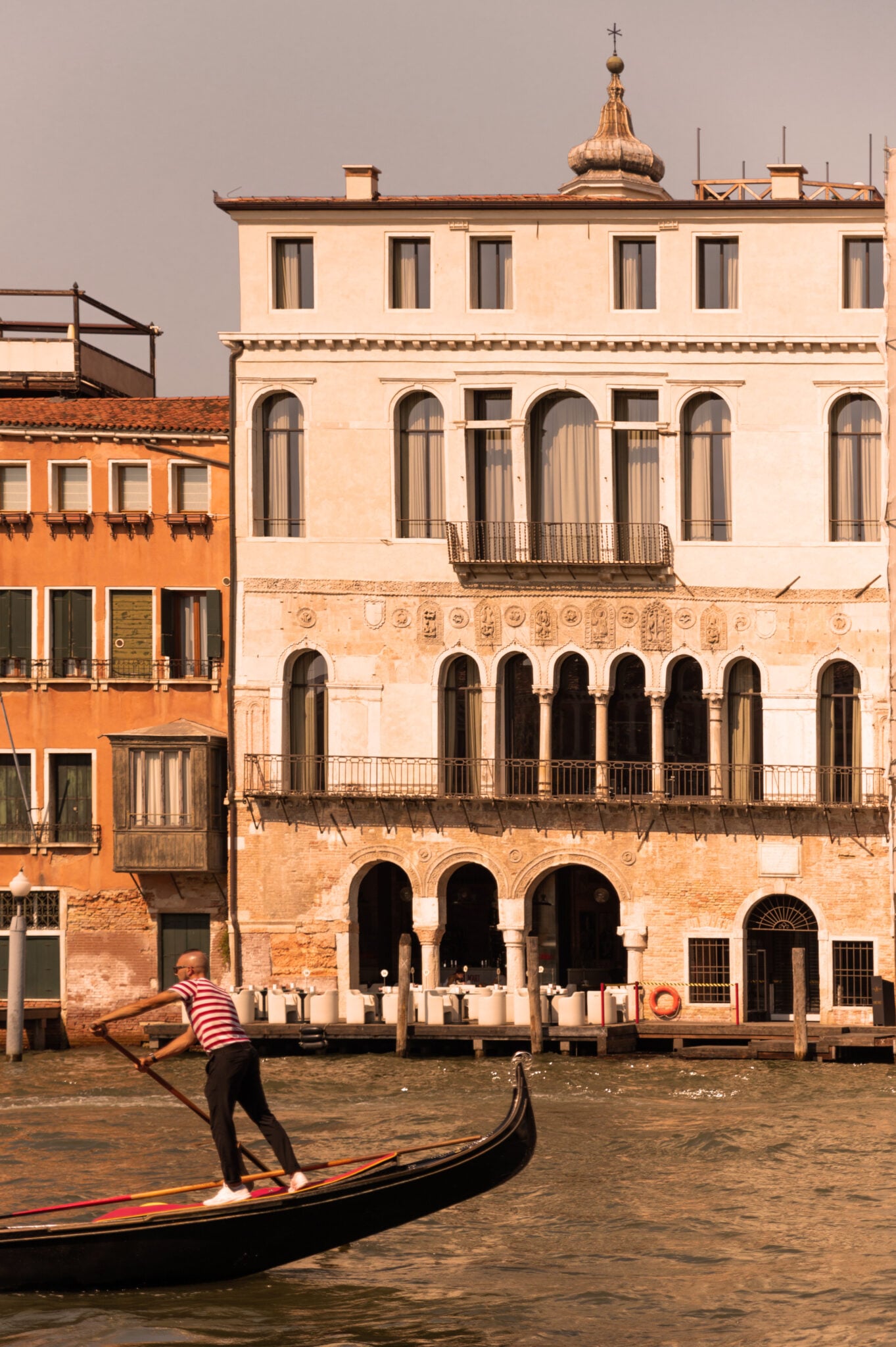 The Venice Venice Hotel, Venice, Italy — Gondolier in striped shirt rowing past terracotta palazzo with arched loggia on Grand Canal at golden hour.