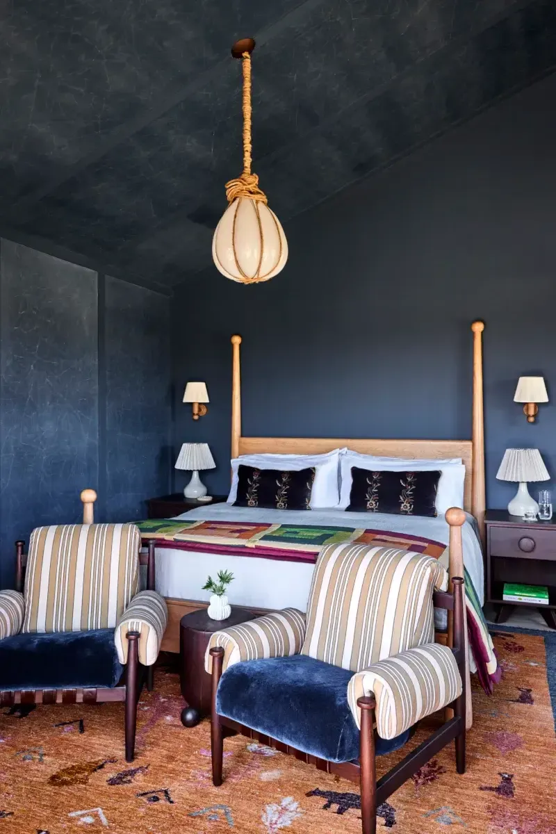 Wildflower Farms, North America — Bedroom with deep blue walls, wooden four-poster bed, striped armchairs, rope pendant light, and colorful patterned rug.
