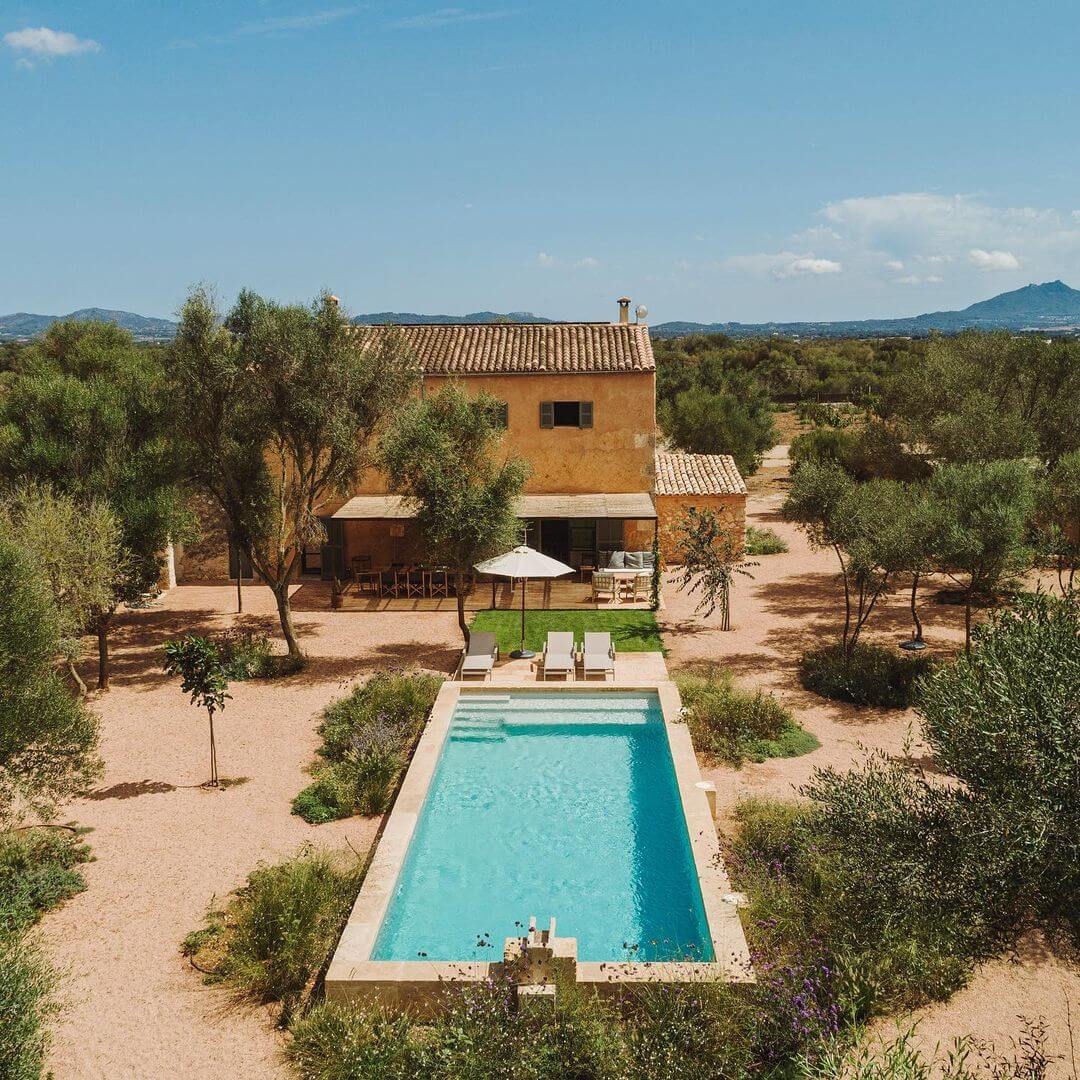 Can Lluïssó, Balearic Islands, Spain — Ochre stone villa with terracotta roof surrounded by olive trees, turquoise lap pool, and loungers on reddish earth courtyard under clear sky.