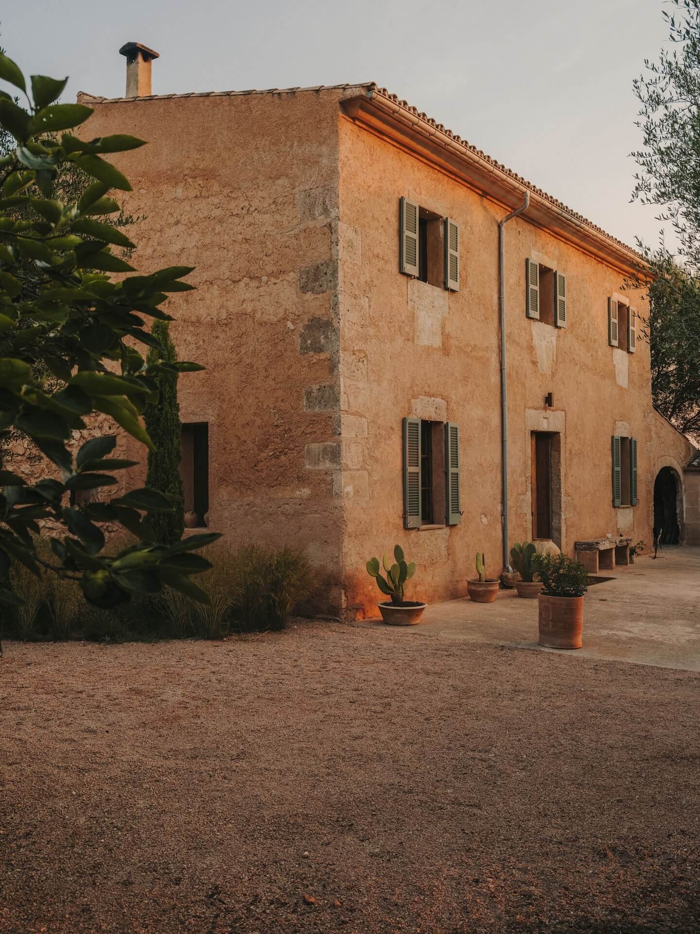 Can Lluïssó, Balearic Islands, Spain — Terracotta stone villa with green shutters, patio courtyard, potted cacti, warm golden-hour light, Mediterranean vernacular architecture.