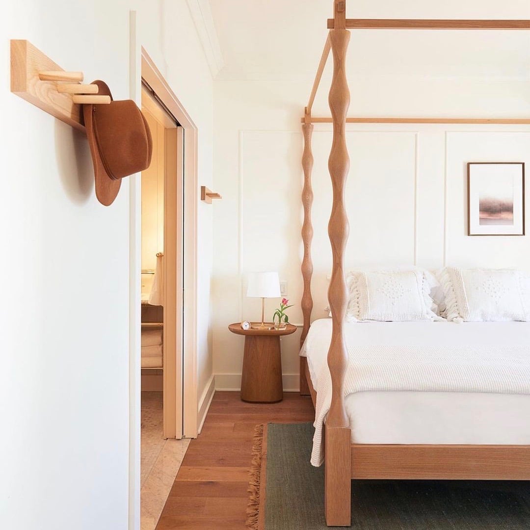 The Lake House, North America — Light wood four-poster bed with twisted posts in minimalist bedroom, natural light, white bedding, wooden nightstand with small plant, framed artwork.
