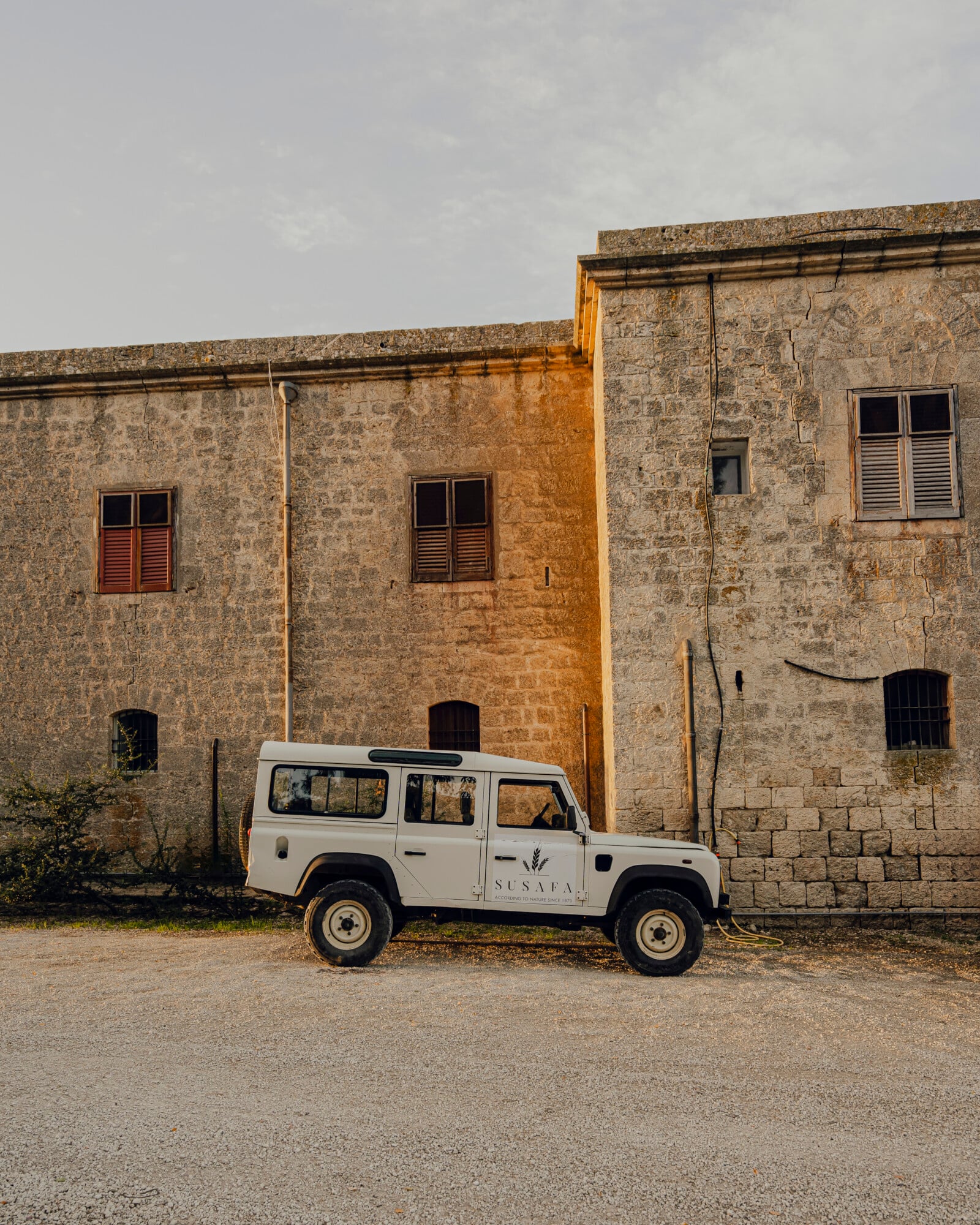 Explore the Region — White Land Rover Defender parked against weathered stone fortress facade with rust-stained walls and wooden shuttered windows, rural Italian countryside.