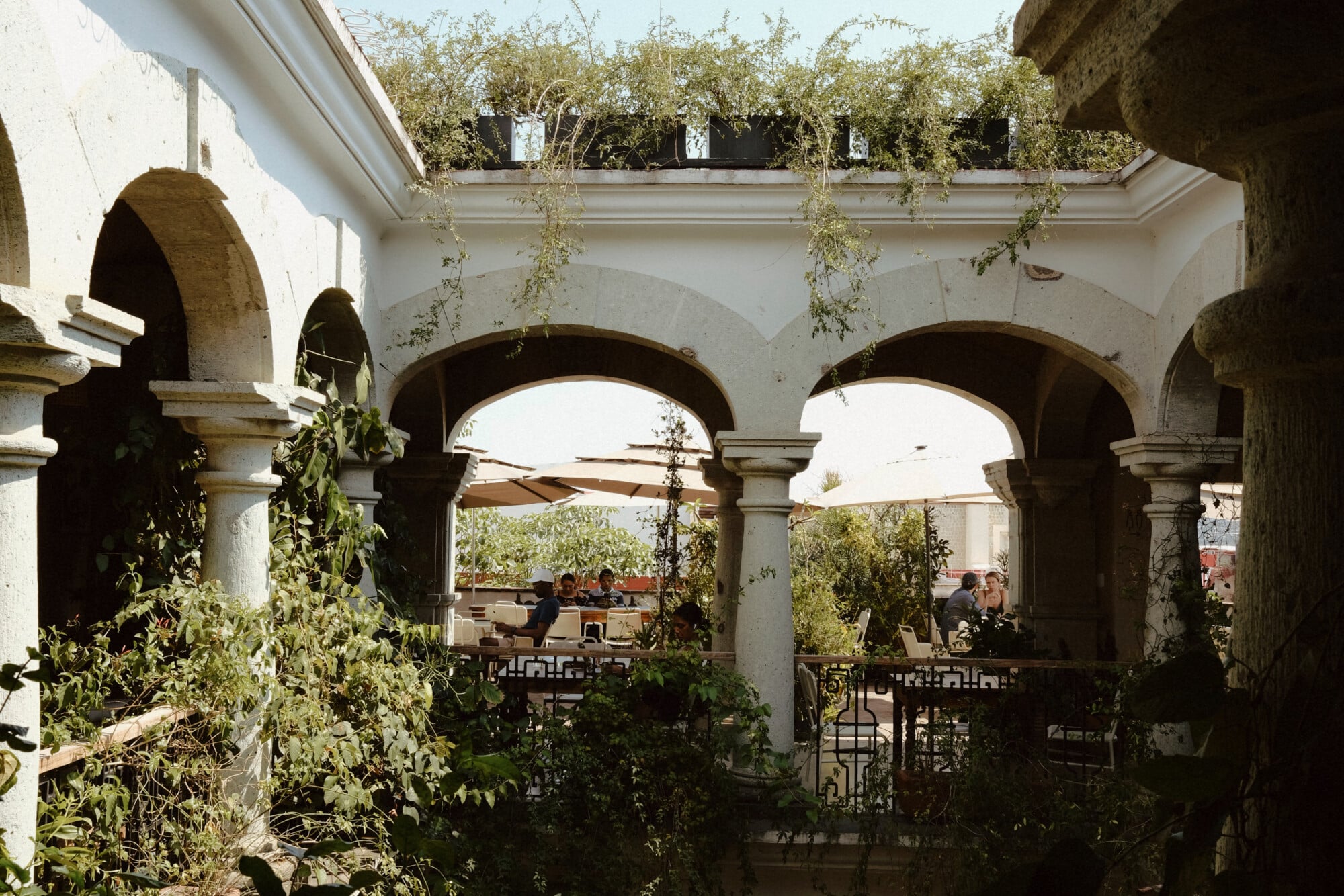 Casa Antonieta, Mexico — Arched stone colonnade with hanging ivy, white walls, and diners visible through open terrace beneath cafe umbrellas.