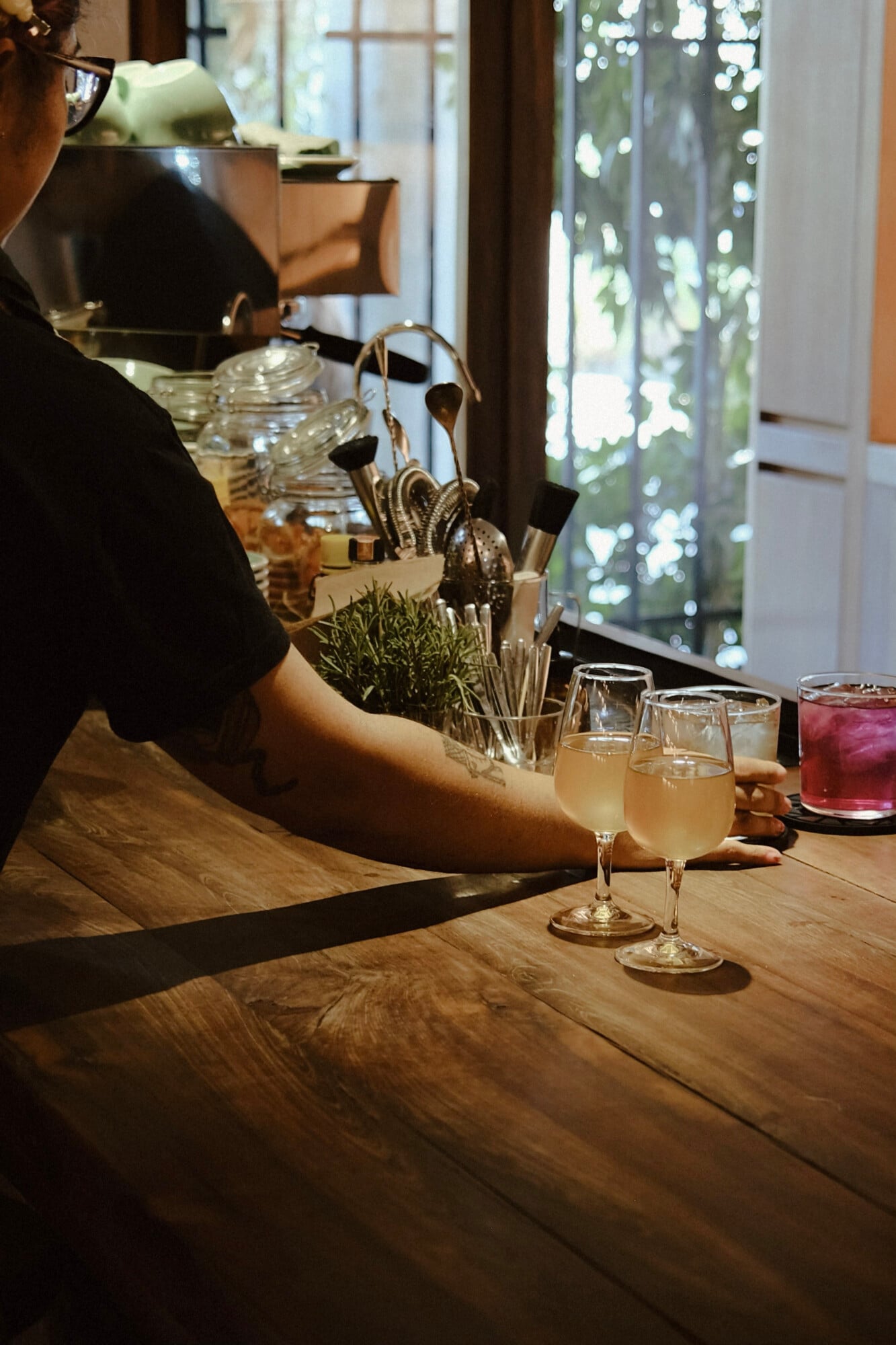 Casa Antonieta, Mexico — Woman at wooden bar with pale cocktails, fresh rosemary garnish, brushed brass fixtures, and garden views through floor-to-ceiling windows.