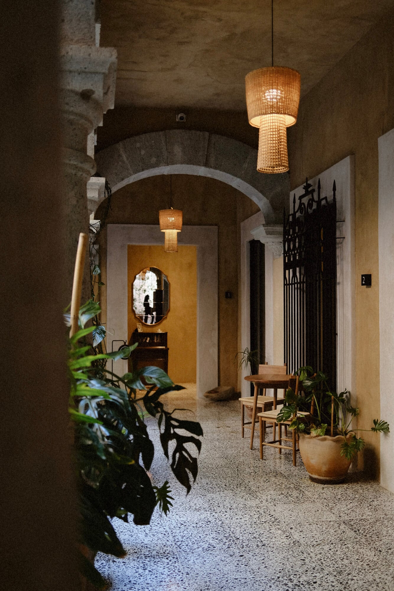 Casa Antonieta, Mexico — Arched corridor with rattan pendant lights, terrazzo flooring, potted plants, wooden chairs, and ochre doorway opening to courtyard light.
